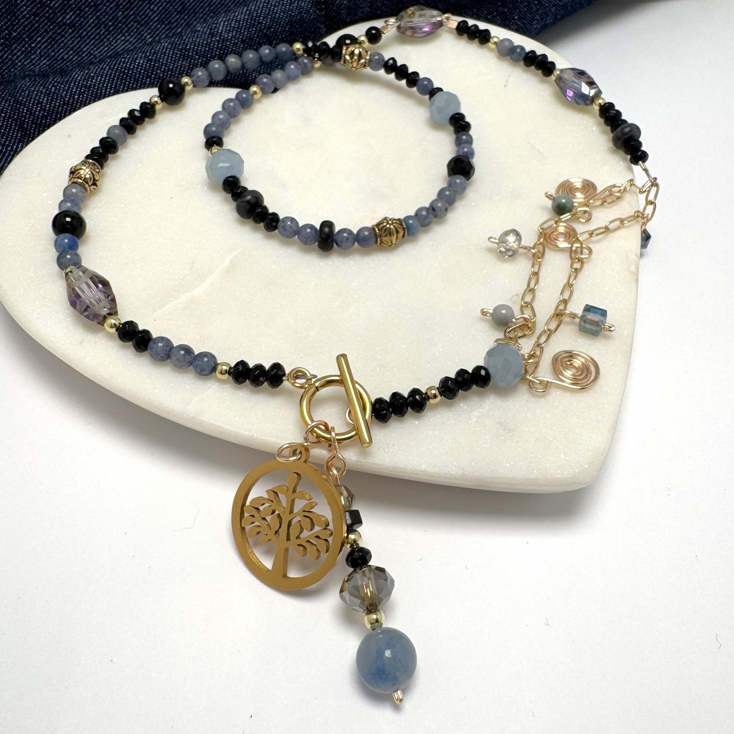 A handcrafted asymmetrical necklace featuring polished blue aventurine and black agate beads, accented with a textured chain and eclectic charms for a modern edge. The toggle clasp is adorned with a gold-toned Tree of Life charm symbolizing strength and connection, along with a unique handmade charm. Perfect for layering or wearing solo, this statement piece by Lacey & Bohème embodies effortless cool and contemporary elegance.