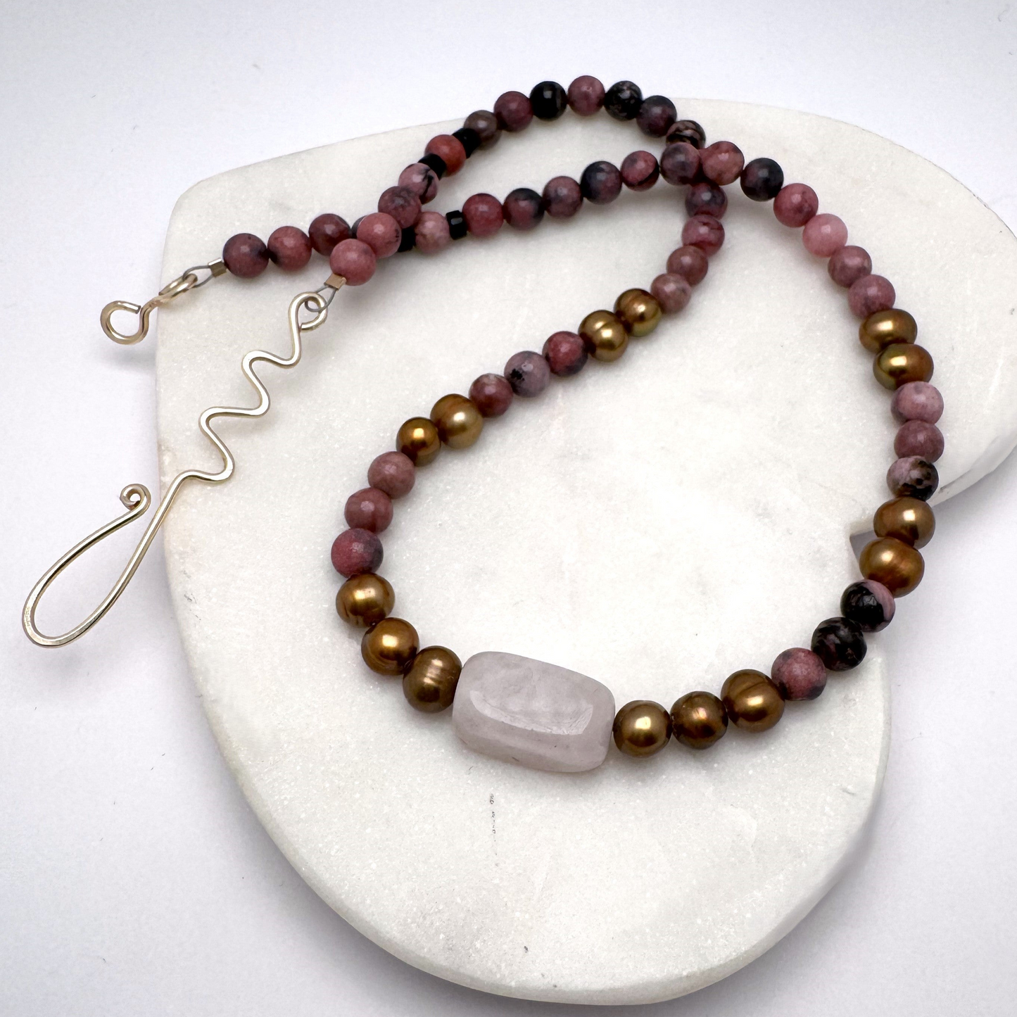 Handmade 19-inch necklace featuring rhodonite, gold-colored pearls, and a rose quartz centerpiece, symbolizing love and serenity. Finished with a gold-tone hand-forged wire hook clasp for a unique artisan touch.