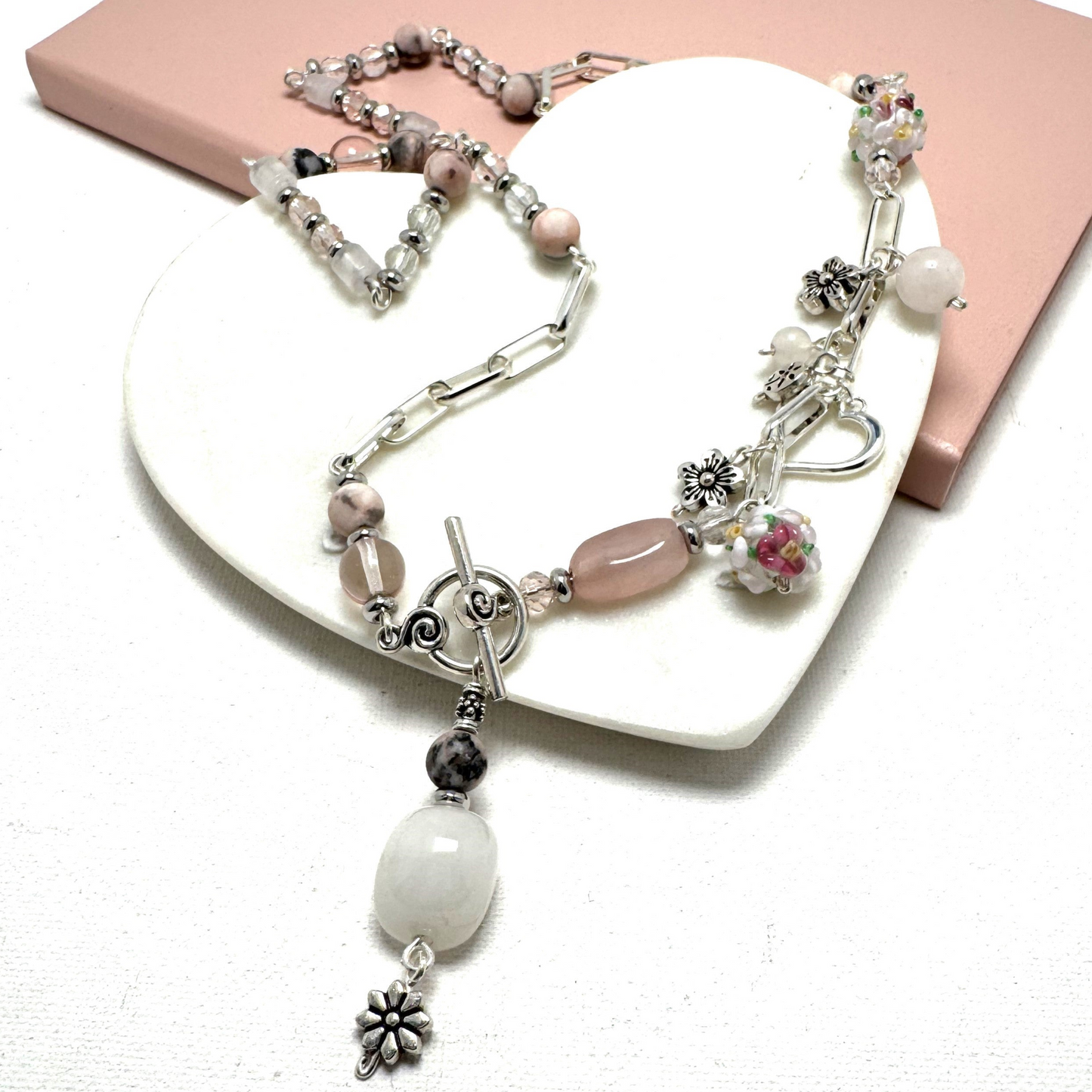 Handmade Blush Harmony Necklace featuring rose quartz, rhodonite, and glass beads with a silver paperclip chain. Accented with a floral lamp work bead, a silver heart, and a front toggle closure with a rose quartz pendant. A romantic and versatile statement piece.