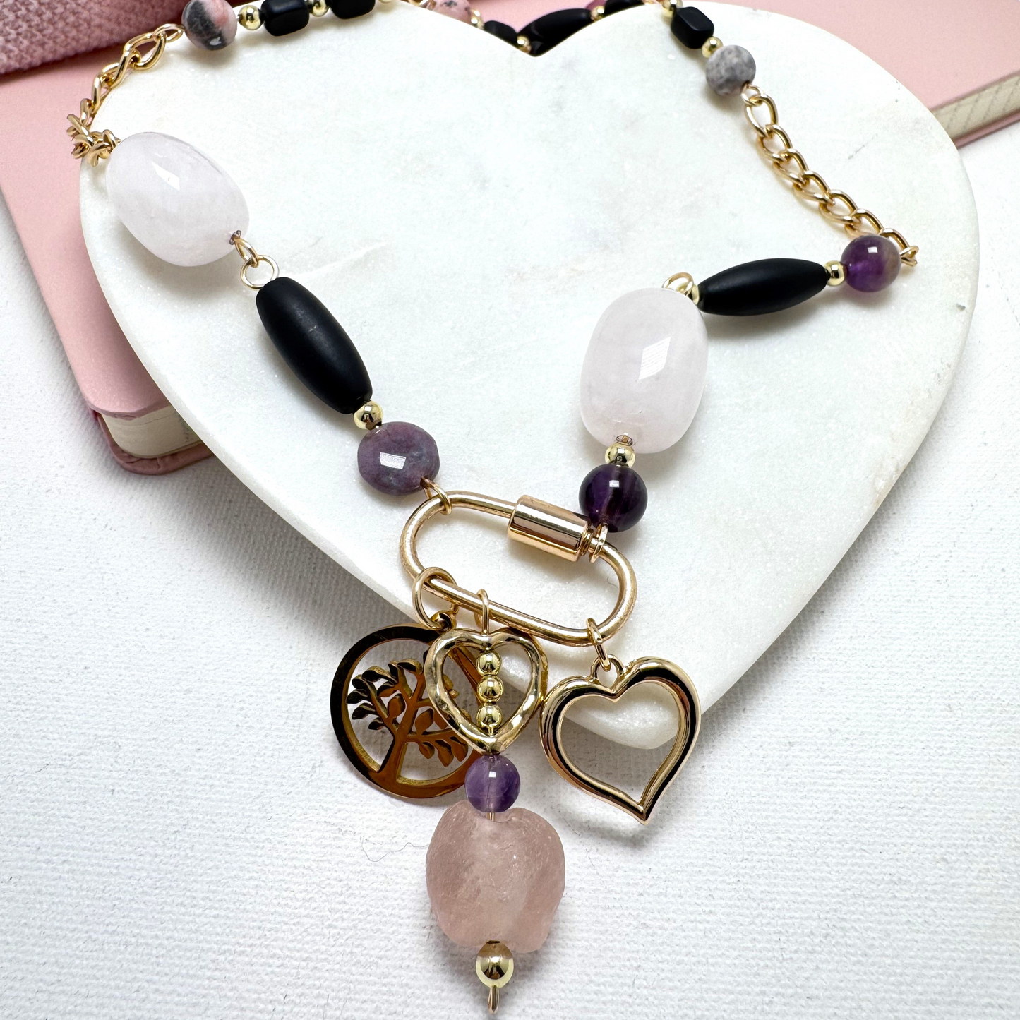 A harmonious blend of organic beauty and contemporary design, this necklace features rose quartz, amethyst, and agate beads intertwined with delicate chain elements. A stylish carabiner serves as the focal point, adorned with three symbolic charms: a Tree of Life, a heart, and pink sea glass with a gold heart. Thoughtfully handcrafted for a meaningful and versatile statement piece.