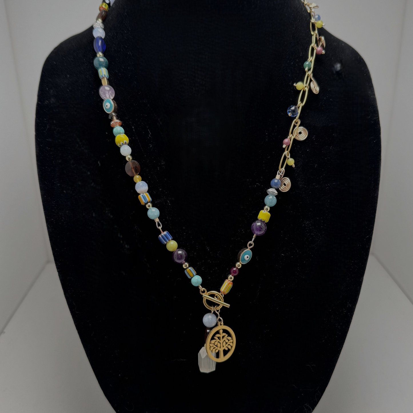 A handcrafted necklace featuring semi-precious stones, ceramic accents, and glass beads, with a modern paperclip chain and delicate stone charms. Anchored by a handmade charm with a trio of beads and a symbolic Tree of Life pendant, this 23-inch necklace by Lacey & Bohème embodies connection, growth, and artisanal beauty. Perfect for everyday wear or special occasions.