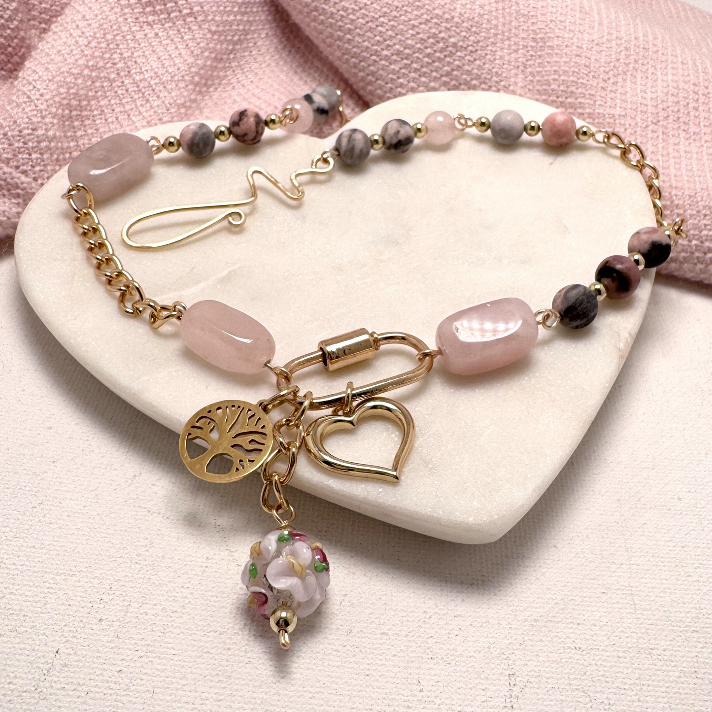 A bold yet feminine Blush Edge Charm Necklace featuring rhodonite and rose quartz stones with a gold chain. The asymmetrical design is centered around a gold carabiner holding three meaningful charms: a Tree of Life, a delicate heart, and a vibrant floral lamp work bead. Finished with a handcrafted forged hook clasp for an artisan touch.