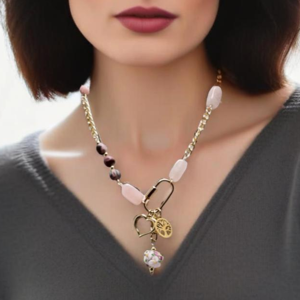 A bold yet feminine Blush Edge Charm Necklace featuring rhodonite and rose quartz stones with a gold chain. The asymmetrical design is centered around a gold carabiner holding three meaningful charms: a Tree of Life, a delicate heart, and a vibrant floral lampwork bead. Finished with a handcrafted forged hook clasp for an artisan touch.