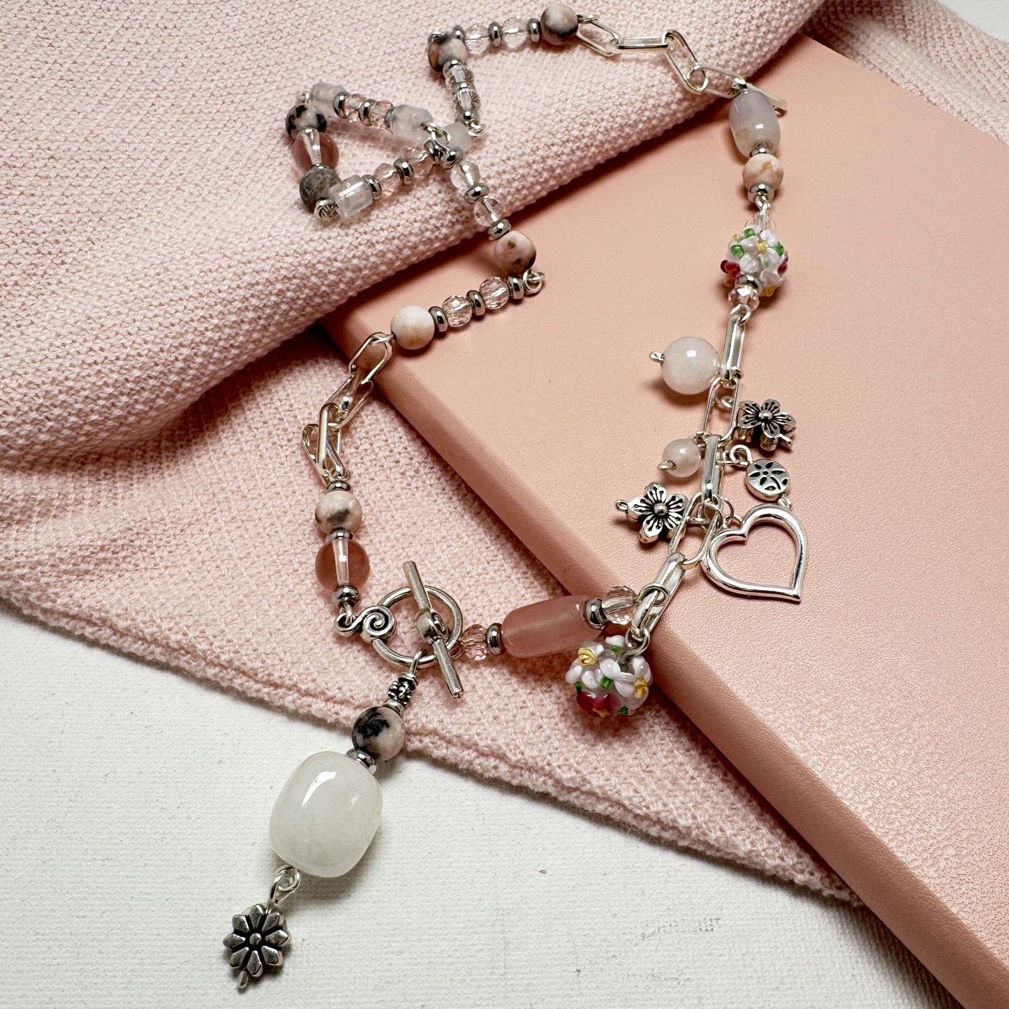 Handmade Blush Harmony Necklace featuring rose quartz, rhodonite, and glass beads with a silver paperclip chain. Accented with a floral lamp work bead, a silver heart, and a front toggle closure with a rose quartz pendant. A romantic and versatile statement piece.