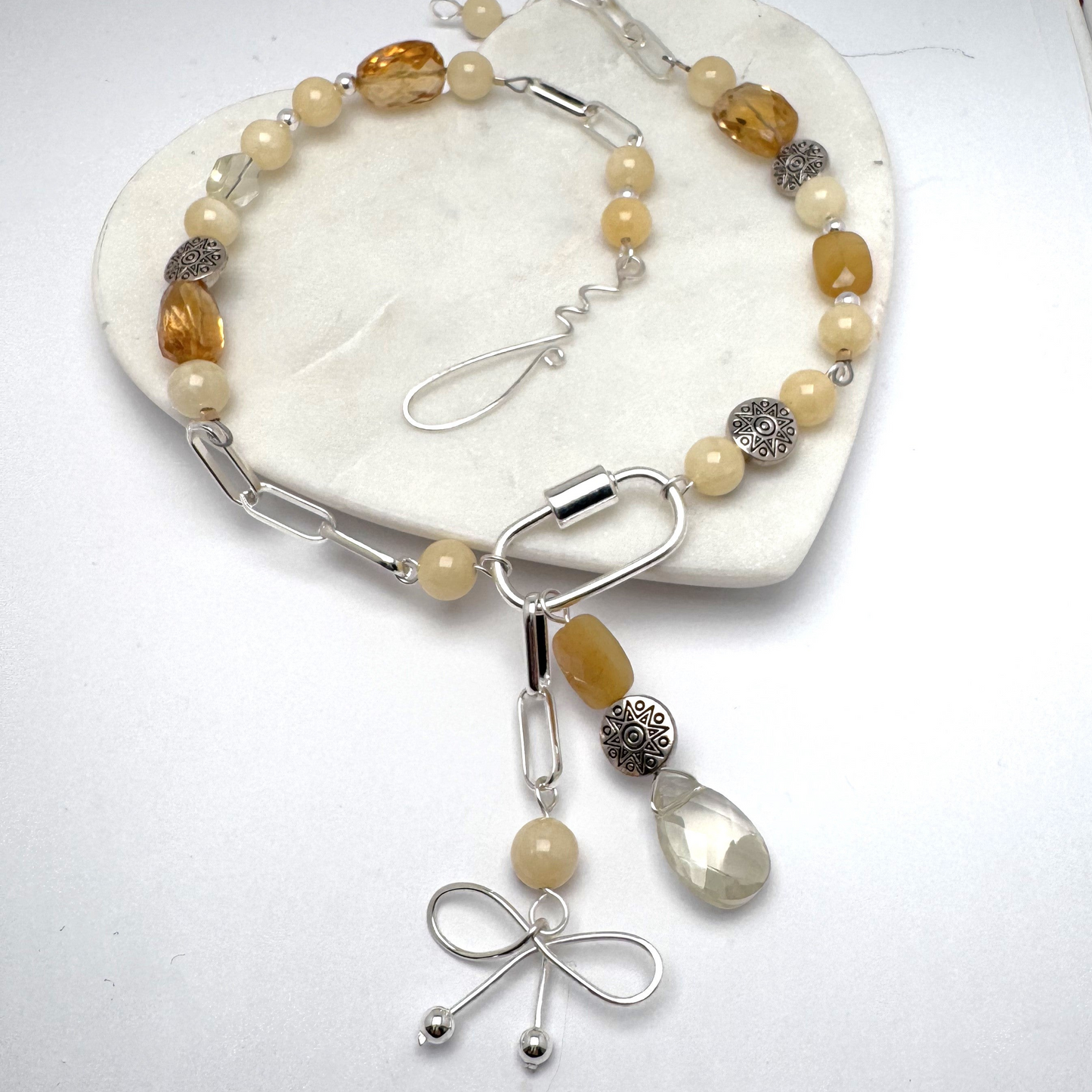 A modern asymmetric necklace featuring a sleek silver chain, vibrant citrine beads, and a silver carabiner clasp holding two unique pendants—a playful bow charm and a mix of citrine beads with a textured silver accent. Finished with a handmade hook clasp, this bold statement piece combines style with citrine’s uplifting energy.