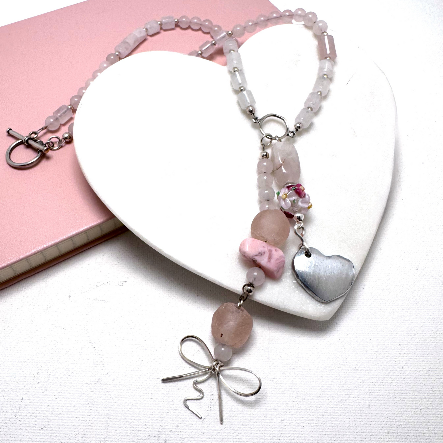 The Blush Bloom Y Necklace is a delicate, handcrafted piece featuring rose quartz, pink opal, sea glass, a floral lamp work bead, and a recycled aluminum heart. Designed with two Y-shaped pendants for a layered look, this 17.5-inch necklace embodies romance and sustainable elegance. Handmade in Canada.