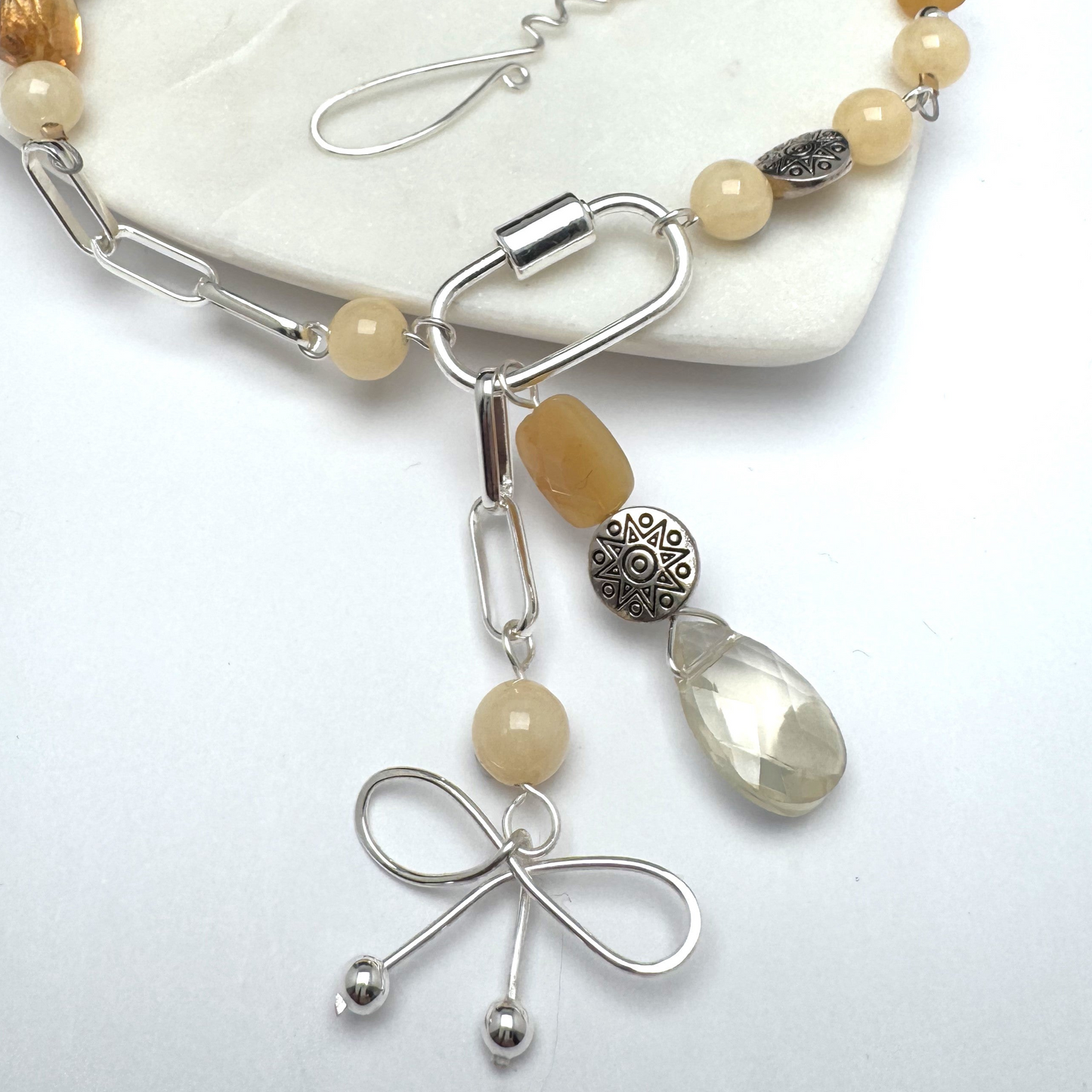 A modern asymmetric necklace featuring a sleek silver chain, vibrant citrine beads, and a silver carabiner clasp holding two unique pendants—a playful bow charm and a mix of citrine beads with a textured silver accent. Finished with a handmade hook clasp, this bold statement piece combines style with citrine’s uplifting energy.