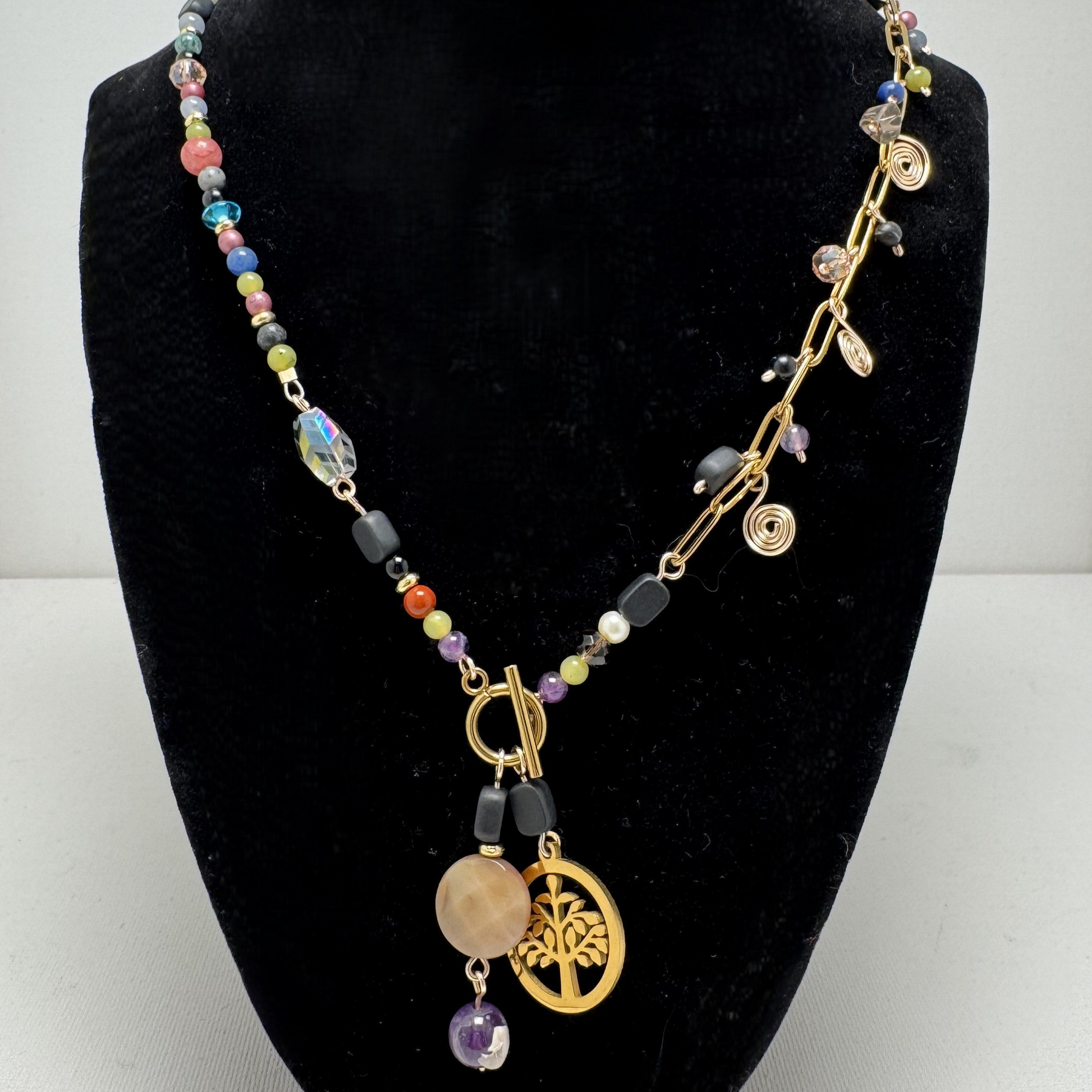 A handcrafted necklace featuring semi-precious stones, ceramic accents, and glass beads, with a modern paperclip chain and delicate stone charms. Anchored by a handmade charm with a trio of beads and a symbolic Tree of Life pendant, this 23-inch necklace by Lacey & Bohème embodies connection, growth, and artisanal beauty. Perfect for everyday wear or special occasions.