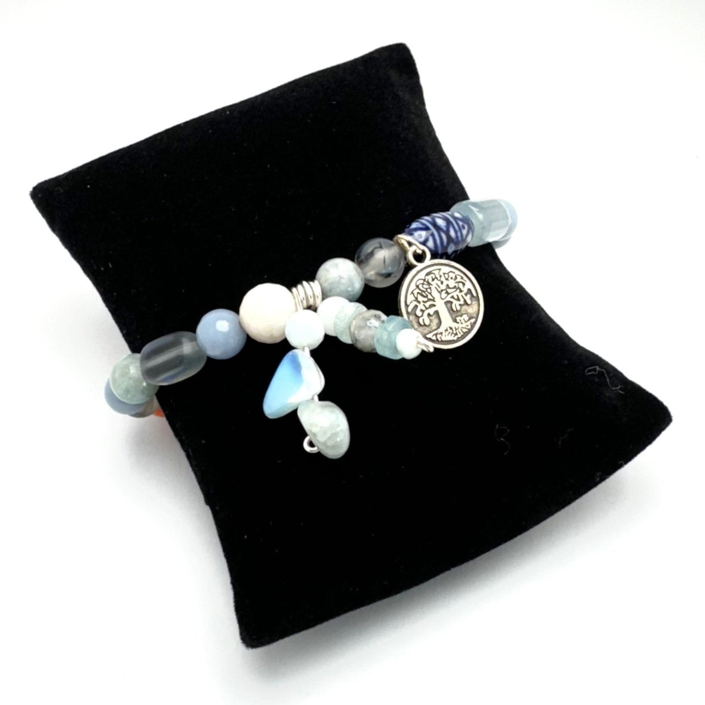 A bracelet with a mix of aquamarine, angelite, glass, and ceramic stones, featuring a tree of life charm, displayed on a black stand.