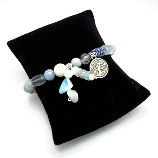 A bracelet with a mix of aquamarine, angelite, glass, and ceramic stones, featuring a tree of life charm, displayed on a black stand.