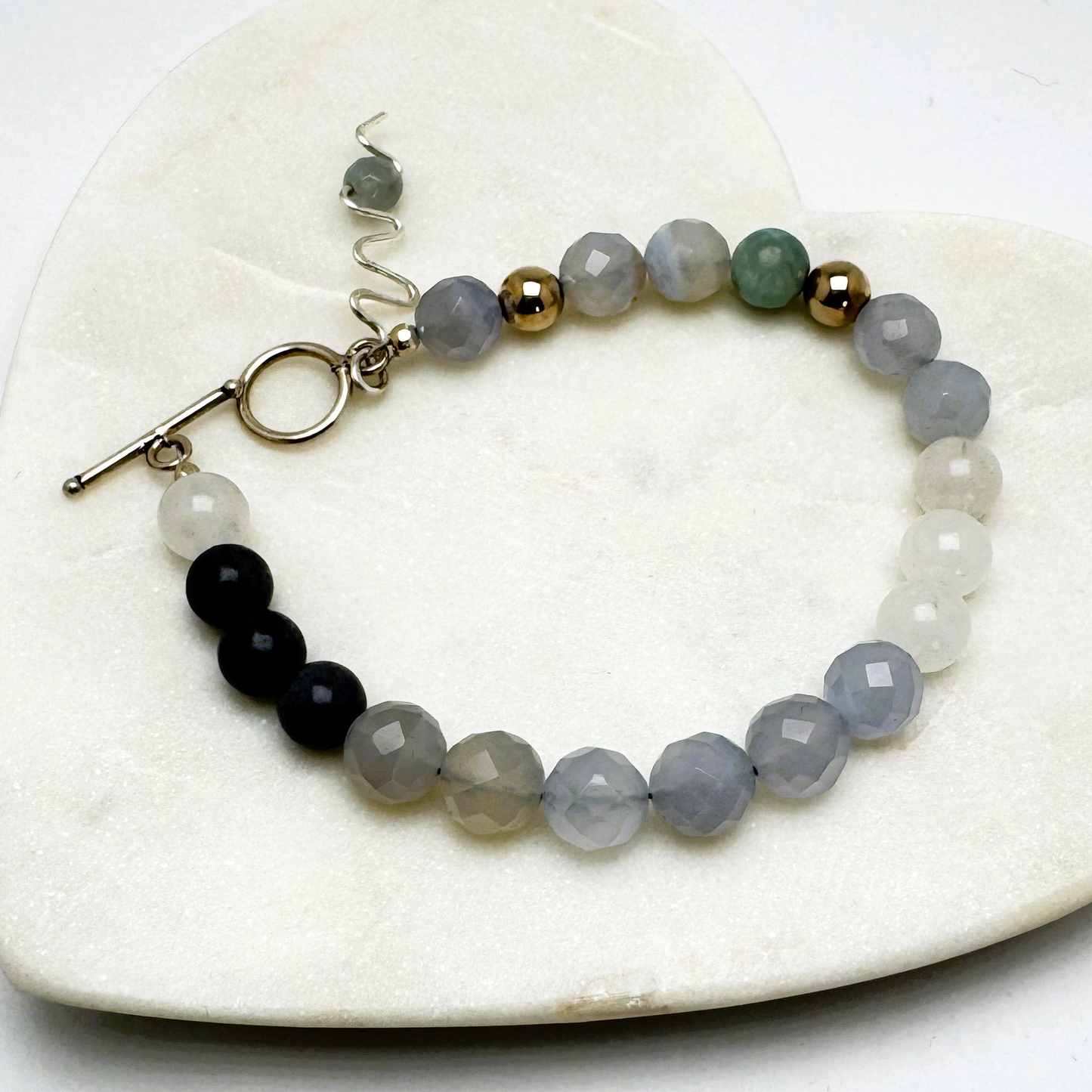 A bracelet with blue chalcedony and aventurine beads, featuring a toggle clasp and a handmade wire charm, displayed on a heart-shaped white stone.