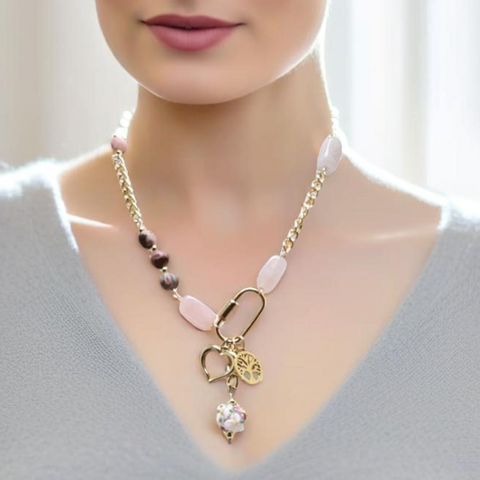 A bold yet feminine Blush Edge Charm Necklace featuring rhodonite and rose quartz stones with a gold chain. The asymmetrical design is centered around a gold carabiner holding three meaningful charms: a Tree of Life, a delicate heart, and a vibrant floral lamp work bead. Finished with a handcrafted forged hook clasp for an artisan touch.