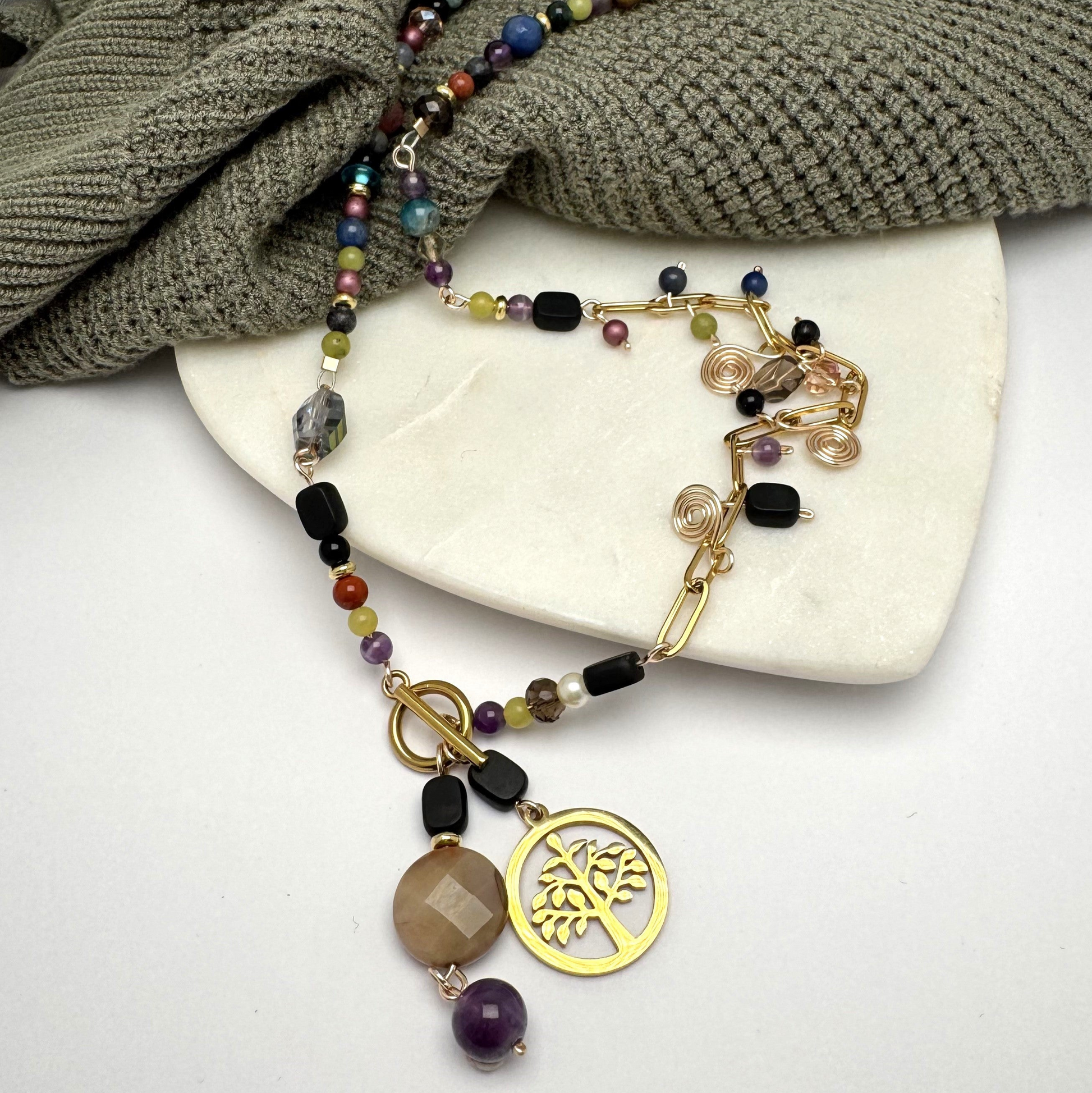 A handcrafted necklace featuring semi-precious stones, ceramic accents, and glass beads, with a modern paperclip chain and delicate stone charms. Anchored by a handmade charm with a trio of beads and a symbolic Tree of Life pendant, this 23-inch necklace by Lacey & Bohème embodies connection, growth, and artisanal beauty. Perfect for everyday wear or special occasions.